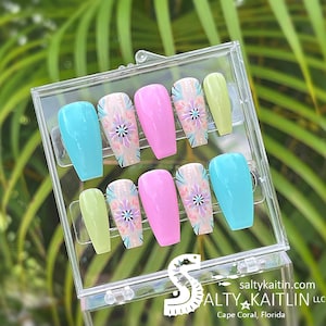 May include: A set of ten false nails in a clear plastic box. The nails are in various colors, including pink, blue, and green. Each nail has a white and purple floral design.