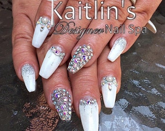 May include: White nail design with glitter and rhinestone accents. Some nails are fully covered in rhinestones, while others have glitter tips or rhinestone borders. The image includes the text "Kaitlin's Designer Nail Spa".
