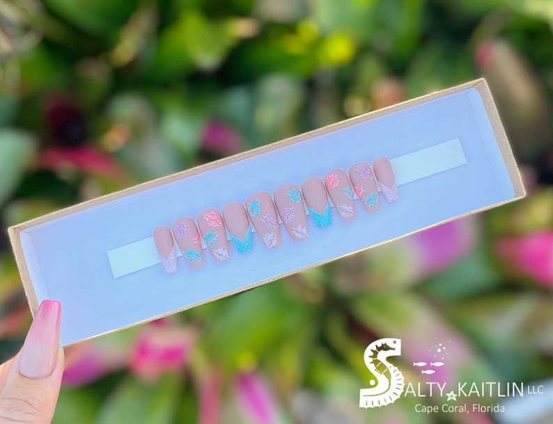 May include: A set of ten false nails with a light pink base and a white tip. Each nail has a different design featuring a small butterfly with glitter accents in shades of pink, blue, and green.