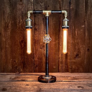 May include: An industrial table lamp with a vintage design. It features a black pipe frame with brass fittings and two elongated Edison-style light bulbs. A valve handle adds a unique touch. The lamp sits on a round base, suitable for a desk or side table.