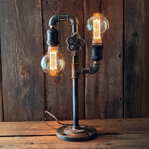 May include: An industrial table lamp constructed from black metal pipes and fittings. It features two Edison-style light bulbs with visible filaments, a valve detail, and a round base. The warm glow of the bulbs contrasts with the dark metal, creating a vintage aesthetic.