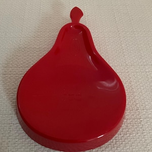 May include: A glossy red plastic tray shaped like a pear. The tray features a smooth, rounded base that tapers upward into a small stem-like protrusion at the top. The surface is reflective and uniform in color, with a slightly recessed center area.
