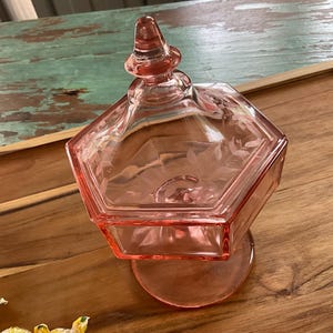 Pode incluir: Uma bomboniere de vidro rosa com corpo hexagonal e tampa com remate cônico. O prato assenta sobre uma base redonda e é exibido em uma bandeja de madeira.