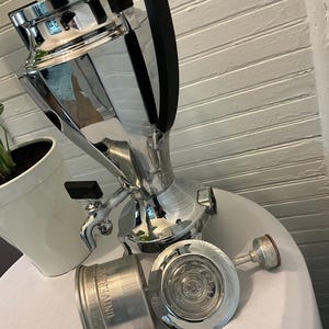 May include: A vintage chrome coffee percolator with a black handle and spigot. The percolator is accompanied by a filter and a glass lid. The word "UNIVERSAL" is visible on the filter. The items are on a white surface.