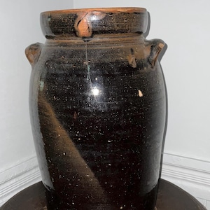 May include: A tall, cylindrical stoneware crock featuring a dark, glossy brown glaze with a visible diagonal streak of lighter brown. The vessel has a thick, reinforced rim, two small side handles, and a prominent vertical crack on the front.