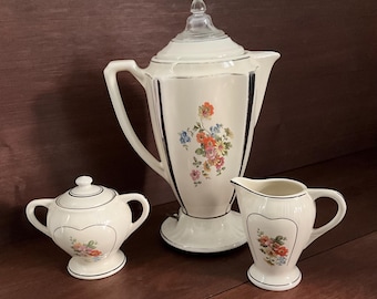 Op de afbeelding: Een crèmekleurig keramisch koffiezetapparaat met glazen deksel, een bijpassende suikerpot met deksel en een melkkannetje. Elk stuk is voorzien van een delicaat bloemmotief met oranje, blauwe en roze bloemen en dunne zilveren randen.