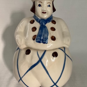 May include: A ceramic cookie jar shaped like a figure wearing a hat, a blue scarf, and a white jacket with brown buttons. The lower portion of the jar features a white base with a blue crisscross pattern and brown circular accents.