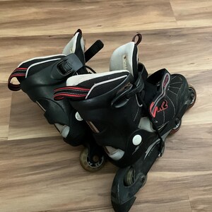 Könnte beinhalten: Paar schwarze Inline-Skates mit roten und weißen Akzenten. Die Skates haben ein hoch geschnittenes Design mit mehreren Riemen und Schnallen für sicheren Halt. Die Rollen sind sichtbar, und das Design konzentriert sich auf Geschwindigkeit und Manövrierfähigkeit.