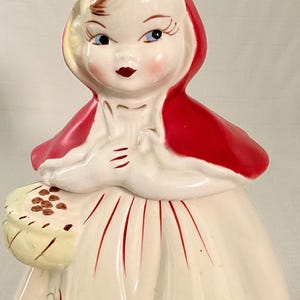 May include: A ceramic figurine of a character wearing a bright red hooded cape over a cream-colored dress. The figure holds a small yellow basket filled with brown items and features painted floral details in red and white along the skirt hem.