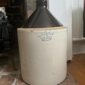 May include: A cylindrical stoneware crock with a cream-colored body and a dark brown glazed shoulder and neck. The front features a blue stamped graphic of a crown with the number 3 inside. A small cork stopper is inserted into the top opening.