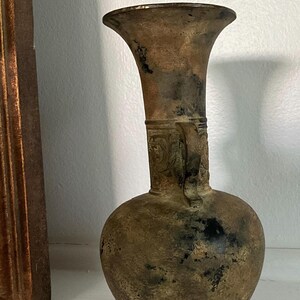 May include: A weathered bronze-toned vase with a bulbous body, a slender neck, and a flared rim. The surface features a mottled patina with dark brown and black spots, and a single handle is attached to the neck, which displays embossed patterns.