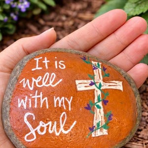 May include: Hand-painted orange stone with the words "It is well with my Soul" in white script. A white cross is adorned with green vines and purple flowers. A unique, inspirational decorative item.