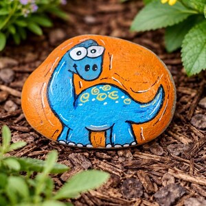 Può includere: Sasso dipinto a mano con un dinosauro blu a fumetti su sfondo arancione. Il dinosauro ha grandi occhi e dettagli gialli. Il sasso è posato su pacciame, circondato da vegetazione.