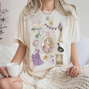 Op de afbeelding: Crèmekleurig T-shirt met cartoonillustraties. Het ontwerp bevat een portret van een blonde vrouw, een toren, een koekenpan, een kroon en andere gerelateerde elementen. Het shirt is een casual kledingstuk.