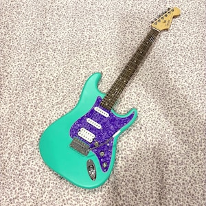 Könnte beinhalten: Eine türkisfarbene E-Gitarre mit einem lila Schlagbrett und weißen Tonabnehmern. Die Gitarre hat einen Hals und eine Kopfplatte aus Holz. Der Hintergrund ist ein Blumenmuster.