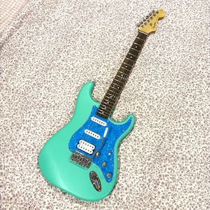 Op de afbeelding: Een turquoise elektrische gitaar met een blauwe, gespikkelde slagplaat. De gitaar heeft witte pickups, een zilveren brug en een lichtgekleurde hals en kop. De achtergrond is een stof met bloemenpatroon.