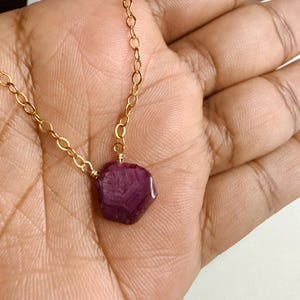 May include: A necklace featuring a raw, hexagonal-shaped deep magenta ruby pendant suspended from a delicate gold-toned metal chain. The gemstone displays a natural, faceted texture with visible internal inclusions and a polished surface finish.