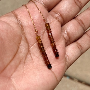 May include: A pair of threader earrings featuring delicate gold-tone chains attached to vertical rows of faceted, translucent amber-colored beads. The beads transition in shade from a lighter golden-yellow at the top to a deep, rich brown at the base.