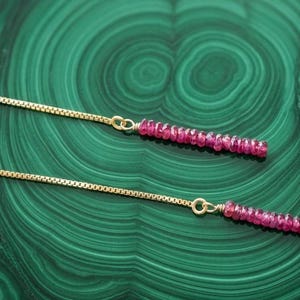 May include: A pair of gold box chain threader earrings, each featuring a dangling linear segment composed of small, faceted, deep pink gemstone beads. The earrings are constructed with a delicate gold chain that connects to the bead strand.