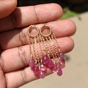 May include: A pair of gold-toned earrings featuring a circular wire-wrapped top connected to multiple delicate gold chains. Each chain terminates in a small, faceted, teardrop-shaped pink gemstone, creating a layered, dangling fringe effect.
