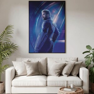 May include: A framed poster featuring a character from a superhero movie. The character is depicted in a dark blue suit, holding a shield. The background is a vibrant blue with light streaks. The poster is hanging above a white sofa with beige pillows.