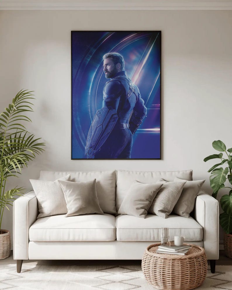 May include: A framed poster featuring a character from a superhero movie. The character is depicted in a dark blue suit, holding a shield. The background is a vibrant blue with light streaks. The poster is hanging above a white sofa with beige pillows.