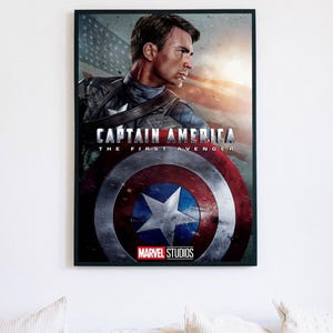 Może przedstawiać: Oprawiony plakat z Kapitanem Ameryką z filmu "Pierwszy Avenger". Obraz przedstawia Kapitana Amerykę z tarczą, amerykańską flagą i logo Marvel Studios. Plakat jest w czarnej ramie i wisi na białej ścianie.