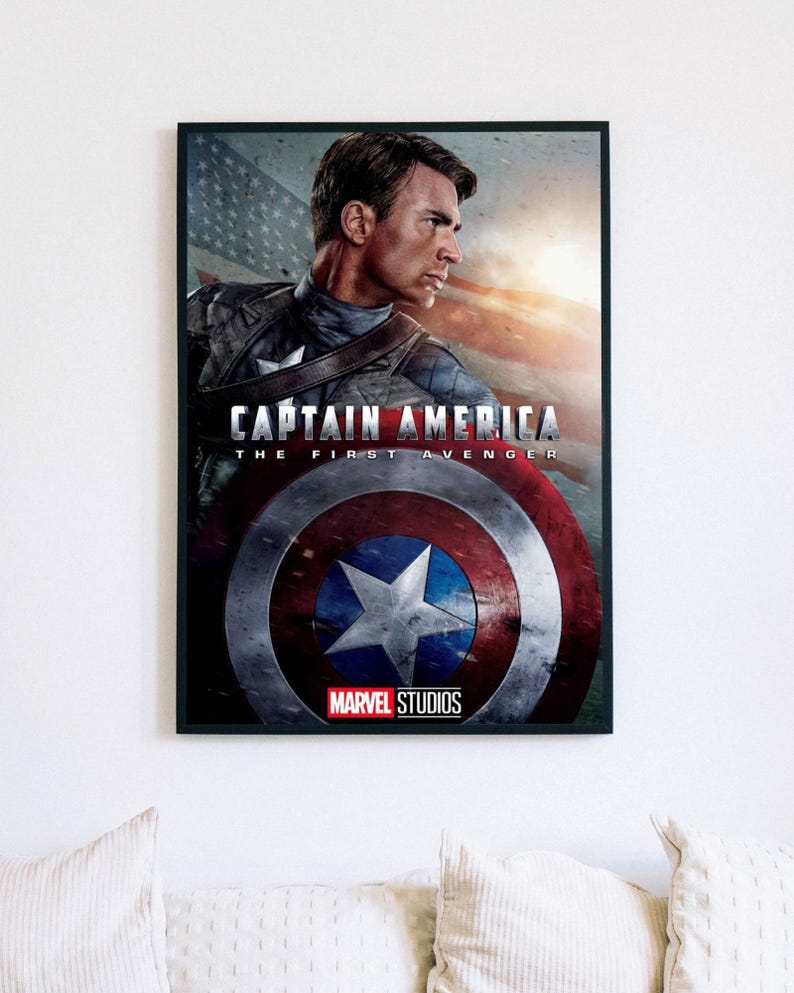 Może przedstawiać: Oprawiony plakat z Kapitanem Ameryką z filmu "Pierwszy Avenger". Obraz przedstawia Kapitana Amerykę z tarczą, amerykańską flagą i logo Marvel Studios. Plakat jest w czarnej ramie i wisi na białej ścianie.