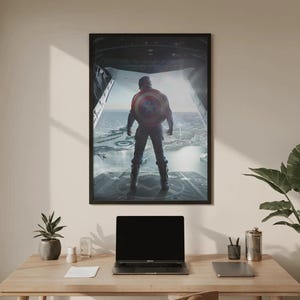 Może przedstawiać: Oprawiony plakat przedstawiający Kapitana Amerykę, stojącego tyłem do widza, z tarczą w dłoni. Grafika przedstawia szczeg&oacute;łową scenę z widocznym w tle krajobrazem miejskim. Plakat jest wyświetlany nad drewnianym biurkiem z laptopem i akcesoriami.