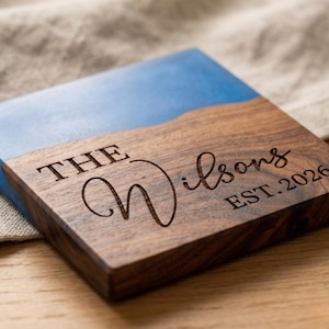 May include: A square wooden coaster with a blue resin top. The coaster is engraved with the words "THE Wilsons EST. 2026". The wood has a rich, dark brown color, and the resin is a vibrant blue.