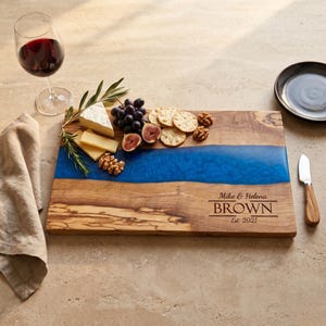 May include: A rectangular wooden serving board featuring a central blue resin river inlay. The board is engraved with the text Mike & Helena BROWN Est 2021. It is displayed with cheese, grapes, figs, walnuts, a wine glass, and a small knife.