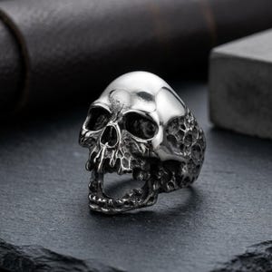 May include: A silver-toned metal ring shaped like a human skull. The skull features a polished, smooth cranium and a highly textured, dark, weathered finish on the jaw and cheekbones, with deep-set eye sockets and an open mouth design.