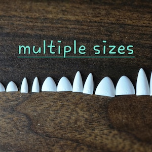 May include: A collection of twelve white, pointed, tooth-like plastic components arranged in a row, varying in width and height. The text "multiple sizes" is printed in light blue above the objects, which feature subtle, molded vertical lines.