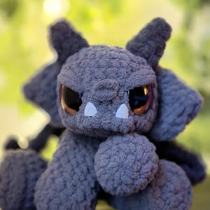 May include: A gray crocheted plush creature with a textured, fuzzy surface. It features large, dark, glossy eyes, two small white triangular teeth protruding from its mouth, and pointed ears. The creature has a rounded head and limbs.