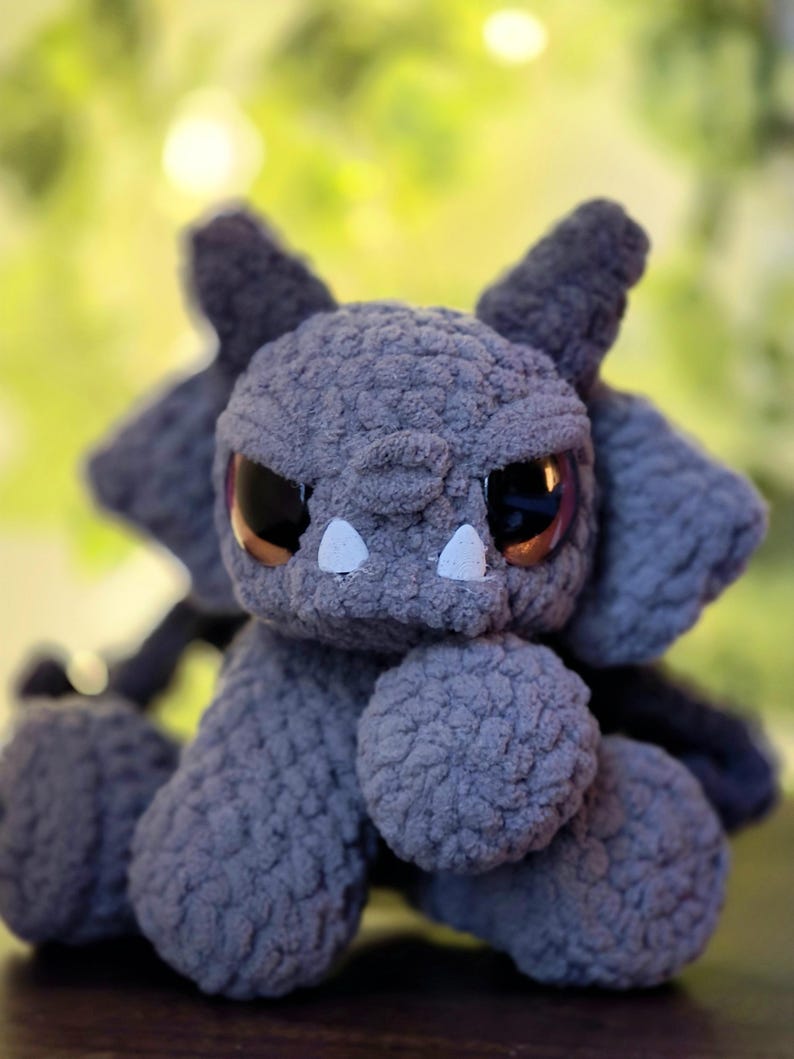 May include: A gray crocheted plush creature with a textured, fuzzy surface. It features large, dark, glossy eyes, two small white triangular teeth protruding from its mouth, and pointed ears. The creature has a rounded head and limbs.