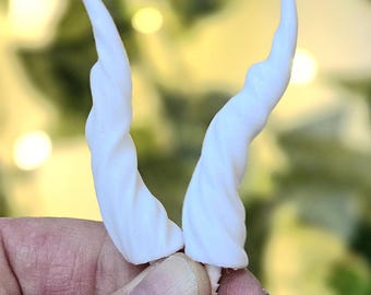 May include: A pair of white, spiraled, tapered horns with a smooth matte finish. The horns curve slightly outward and upward, featuring a distinct twisted texture that runs from the base to the pointed tip of each individual horn piece.