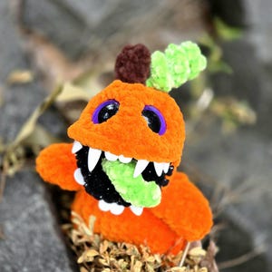 May include: A plush orange creature made of soft yarn with a large open mouth, sharp white teeth, and a bright green tongue. It features dark purple eyes, a brown stem-like top, and a textured green leaf accent on its head.