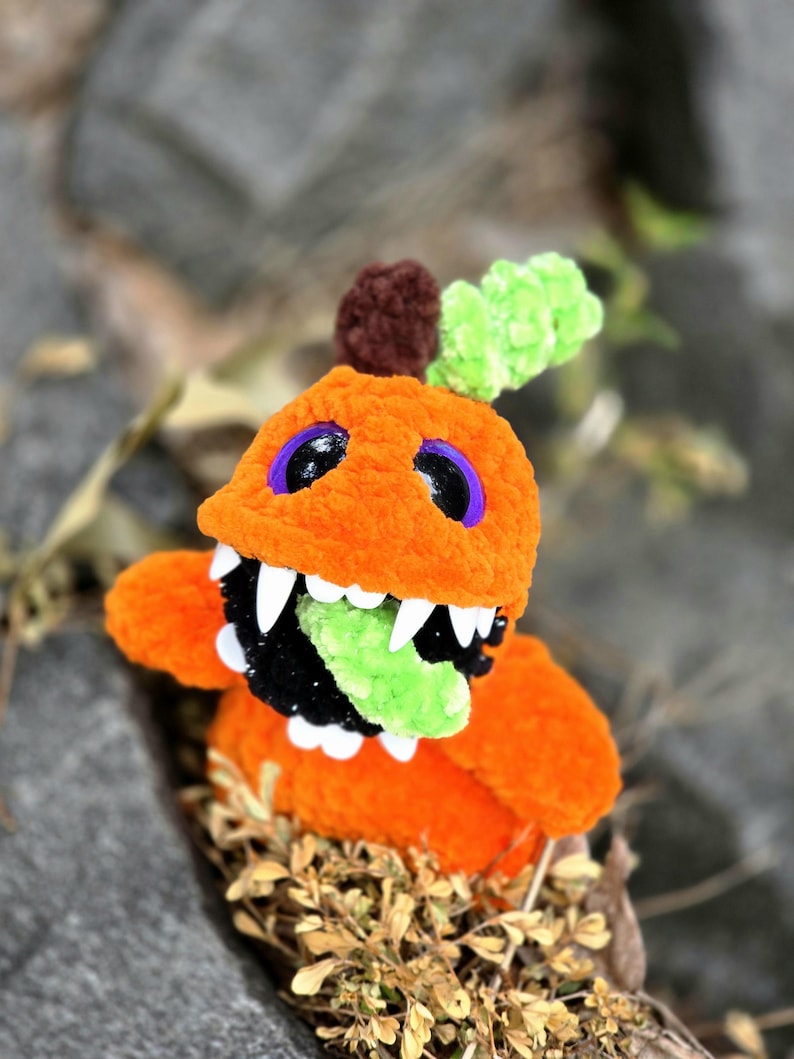 May include: A plush orange creature made of soft yarn with a large open mouth, sharp white teeth, and a bright green tongue. It features dark purple eyes, a brown stem-like top, and a textured green leaf accent on its head.