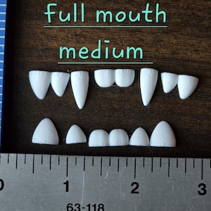May include: A set of white, 3D-printed plastic teeth arranged in two rows, labeled full mouth medium. The teeth feature visible concentric circular textures. A blue plastic ruler and a metal ruler are visible, showing the set is approximately 2.5 inches wide.
