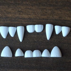 May include: A collection of white, 3D-printed plastic teeth arranged in two rows. The top row features pointed fangs and rounded incisors, while the bottom row consists of smaller, rounded teeth. Each piece displays a visible concentric layer texture.