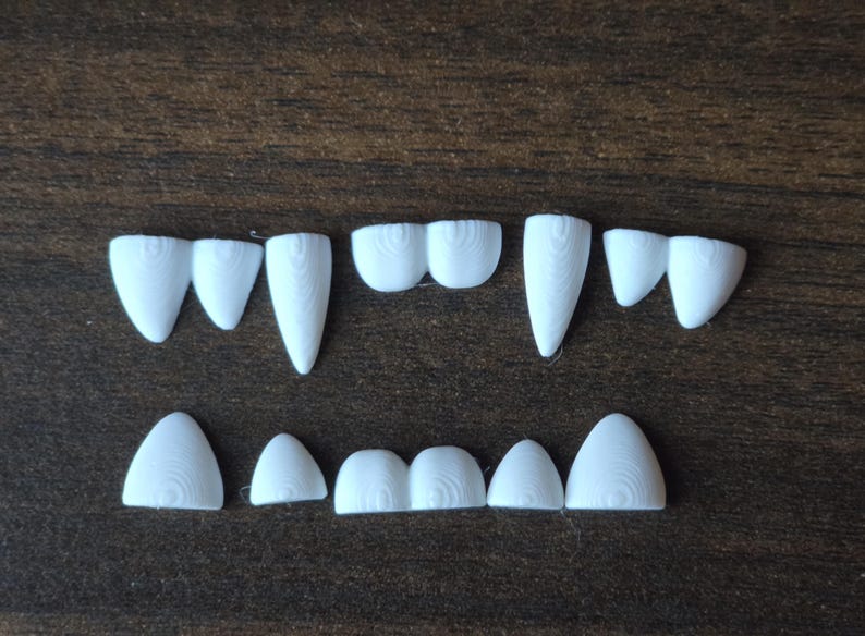 May include: A collection of white, 3D-printed plastic teeth arranged in two rows. The top row features pointed fangs and rounded incisors, while the bottom row consists of smaller, rounded teeth. Each piece displays a visible concentric layer texture.
