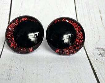 May include: A pair of round, domed cabochons featuring a solid black center surrounded by a vibrant red, glittery iris-like pattern. These circular pieces have a smooth, glossy finish that reflects light, creating a high-contrast, eye-like appearance.
