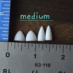 May include: Four small, white, pointed, 3D-printed plastic components arranged in a row. The two on the left are wider and shorter, while the two on the right are narrower and taller. A metal ruler shows the scale in inches, and the word medium is printed above the items.