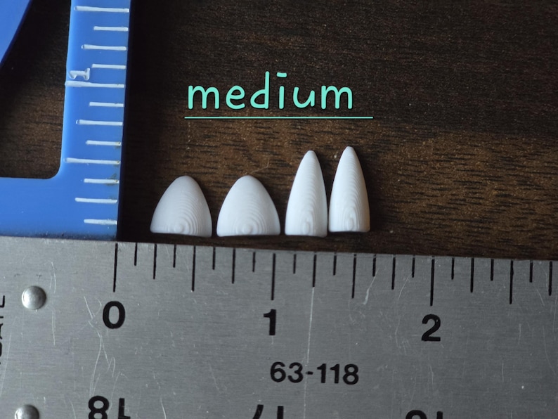 May include: Four small, white, pointed, 3D-printed plastic components arranged in a row. The two on the left are wider and shorter, while the two on the right are narrower and taller. A metal ruler shows the scale in inches, and the word medium is printed above the items.