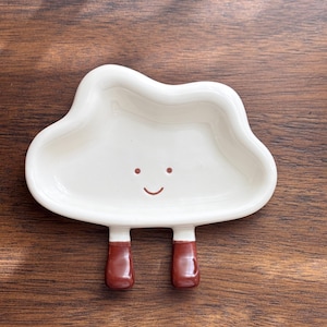 May include: A whimsical, cloud-shaped ceramic dish in off-white with a smiling face. The dish has two brown, rectangular legs. The background is a dark wood surface.