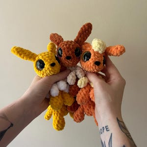 May include: Three small, handmade crochet plush toys made from soft, textured yarn. One is bright yellow, one is dark brown, and one is orange with a cream-colored tuft on its head. Each plush features large, glossy black plastic safety eyes.