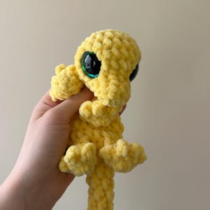 May include: A small, handmade crochet plush toy in a bright yellow color. The creature features a soft, textured yarn construction with large, round, glossy black eyes that have a subtle green shimmer. It has small limbs and a long tail.