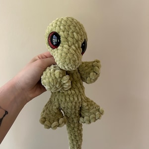 May include: A soft, crocheted plush toy in the shape of a lizard, crafted from light green yarn with a textured, chunky knit appearance. The creature features large, glossy black eyes with a distinct red glittery rim, four small limbs, and a long tail.
