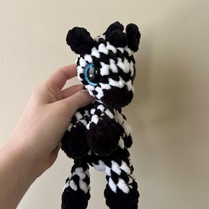 Könnte beinhalten: Ein Plüsch-Zebra aus weichem, dickem Garn mit einem sich wiederholenden schwarz-weißen Karomuster. Das Stofftier hat große, runde, glänzend schwarze Augen mit leuchtend blauen Glitzerrändern, kleine schwarze Ohren und vier ausgeprägte Beine.