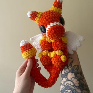 May include: A plush crochet dragon toy featuring a vibrant orange, yellow, and white color scheme reminiscent of candy corn. The creature has large black eyes, small white wings, and a long tail ending in a pointed, multi-colored tip.